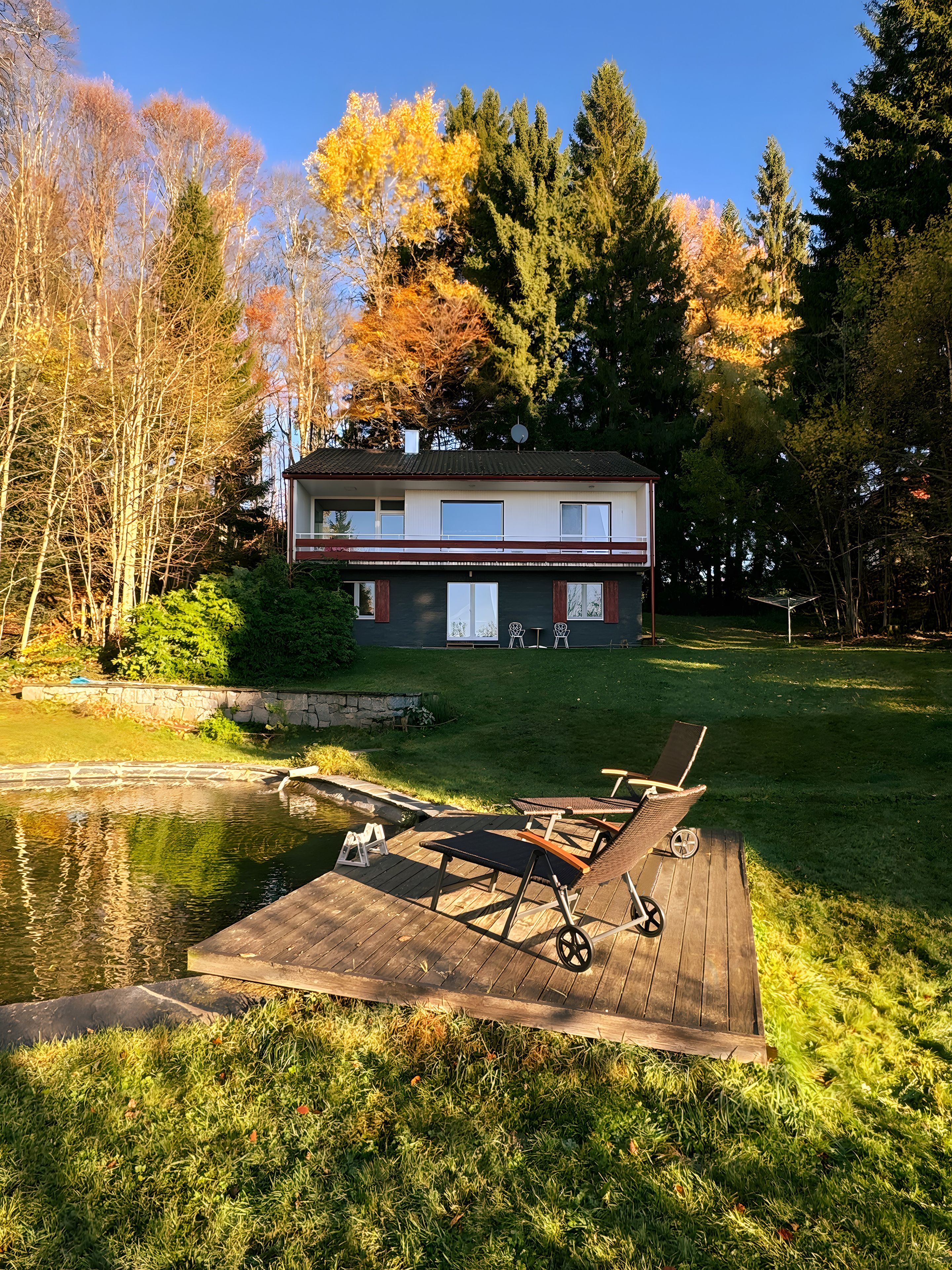 Une maison à deux étages dotée d’une terrasse donnant sur un étang, entourée d’arbres et d’une pelouse verdoyante.