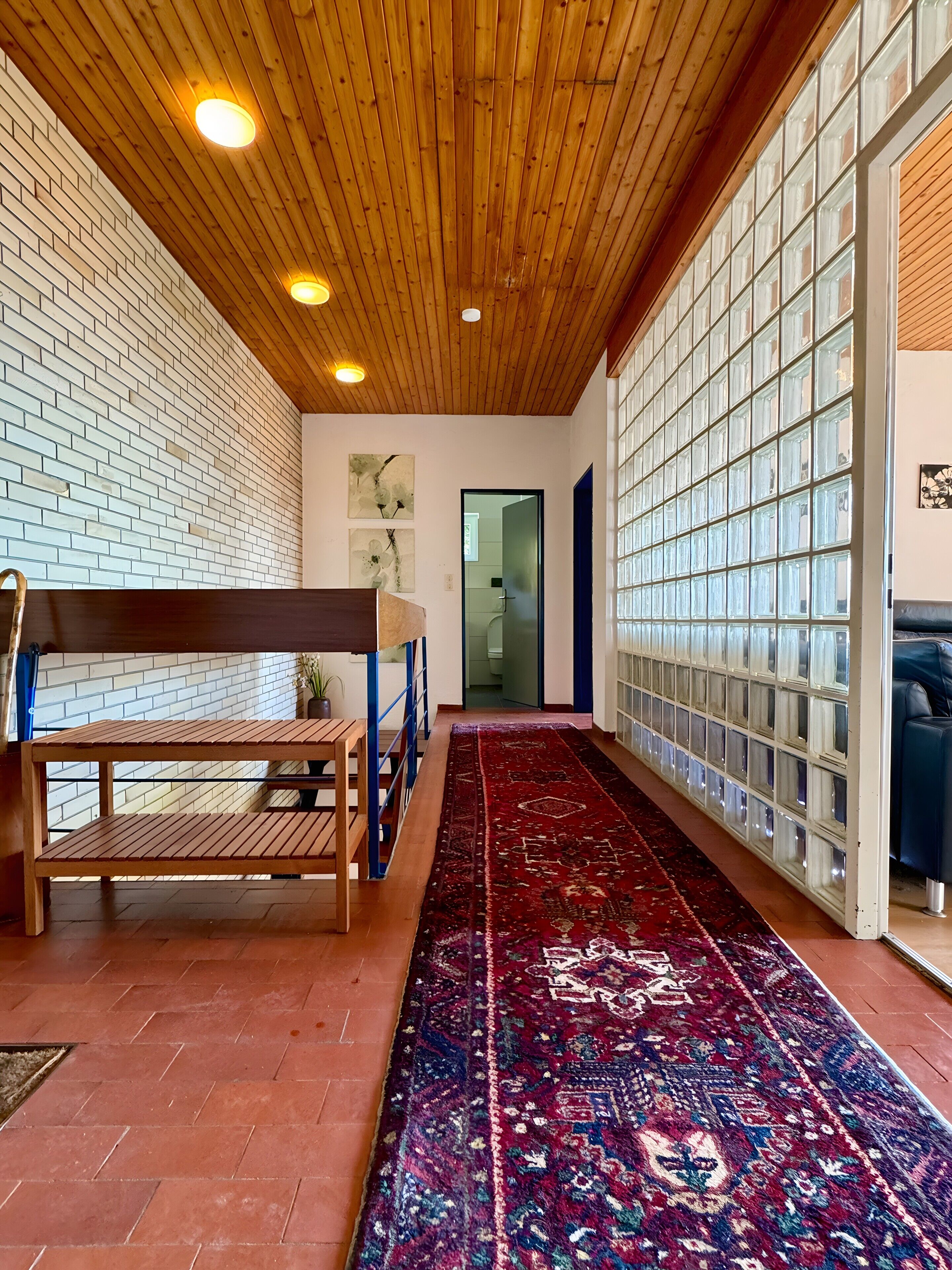 Un couloir avec un plafond en bois, un tapis à motifs, un banc et un mur en blocs de verre.