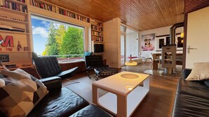 A modern living room with a large window, bookshelves, a sofa, a coffee table, and a fireplace. - Holiday Home 'Alpenblick' with Mountain View, Private Terrace and Wi-Fi (Rickenbach)