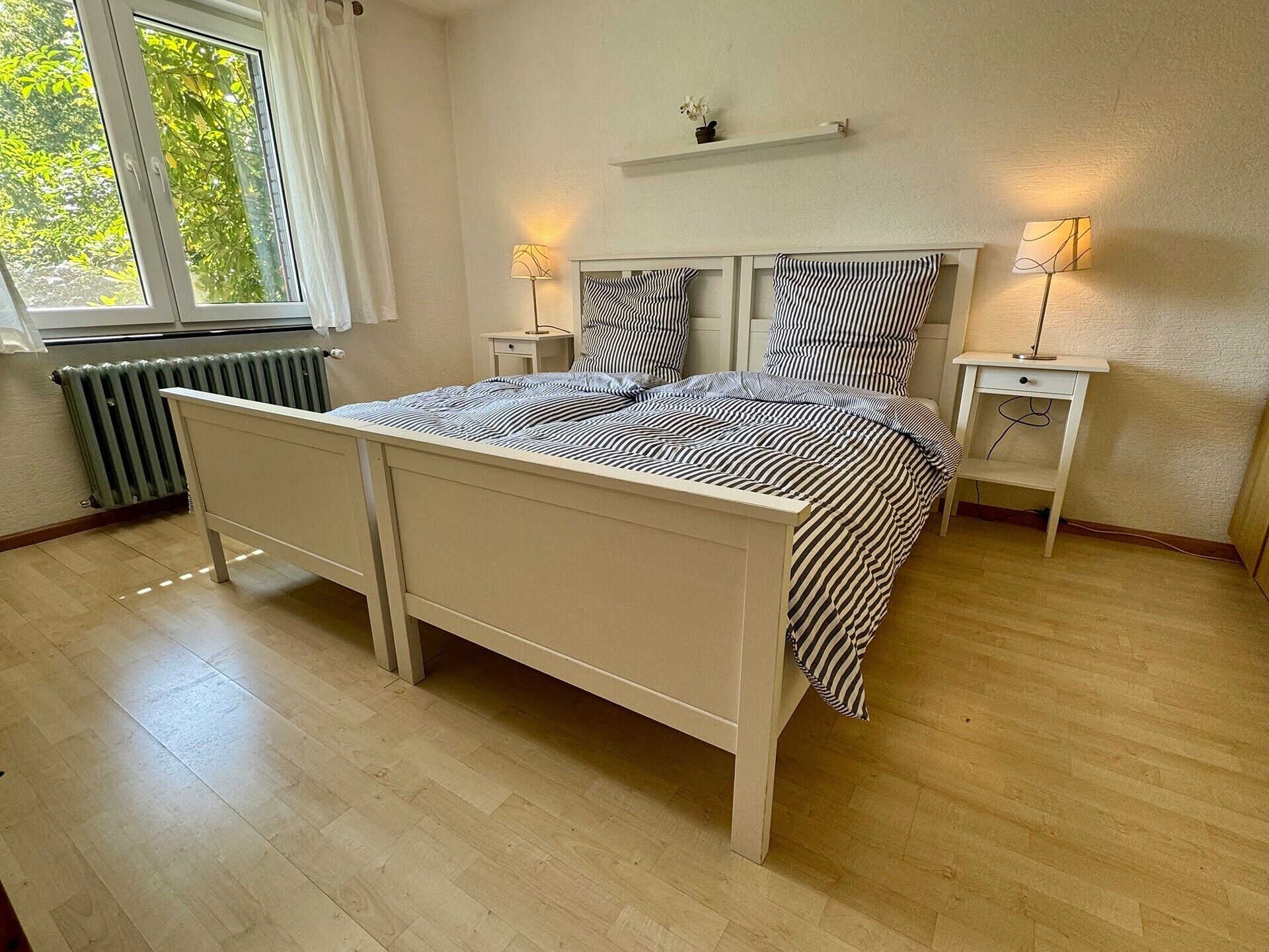A bedroom with a bed, two bedside tables, a radiator, and a window with curtains.