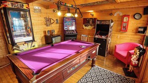 Una mesa de billar en una sala con paredes de madera, juegos de arcade y un sofá rojo.