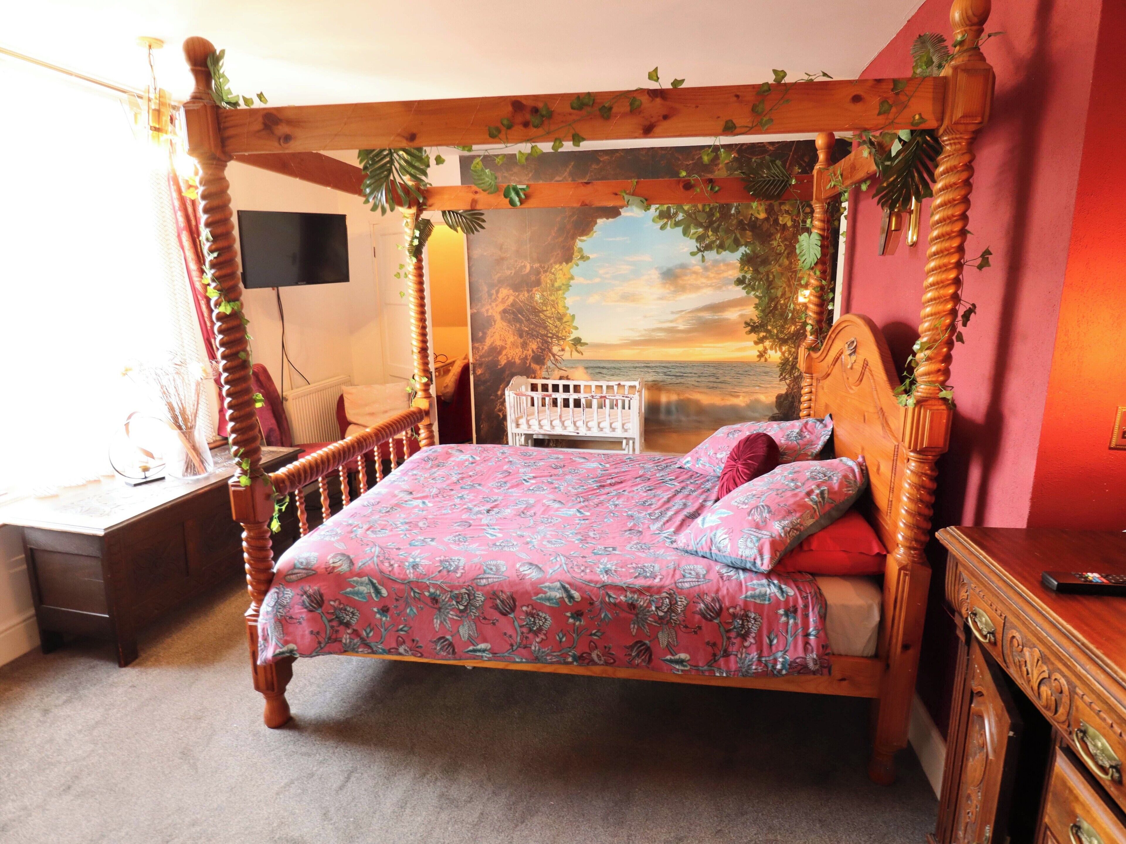 A four-poster bed with a floral bedspread, a white crib, and a wall mural of a sunset.