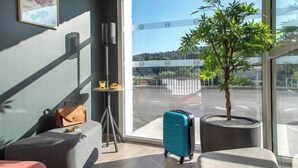 Una habitación de hotel moderna con un ventanal grande, una planta en maceta, una maleta y una pequeña mesa con una bebida.