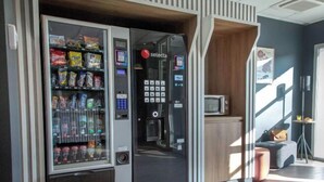 Máquina expendedora con variedad de bocadillos y bebidas, un microondas y una pequeña zona de estar con una mesa y sillas.
