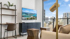 Een moderne woonkamer met uitzicht over de stad, een grote flatscreen televisie, een houten salontafel, een bank en een boekenkast met een plant.