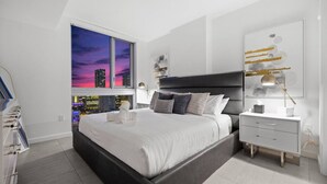Een moderne slaapkamer met een groot bed, een nachtkastje, een bijzettafeltje en uitzicht op de stad bij zonsondergang.
