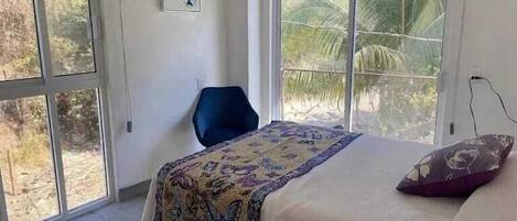 Un dormitori amb un llit, una cadira, una finestra amb vista a palmeres i un quadre a la paret.