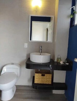 Ein Badezimmer mit einer weißen Toilette, einem weißen Waschbecken mit schwarzer Arbeitsplatte und einem Spiegel darüber. Unter der Arbeitsplatte befindet sich eine kleine Holzkiste.