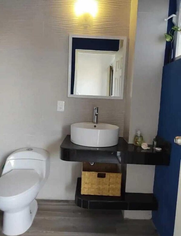 A bathroom with a white toilet, a white sink with a black countertop, and a mirror above the sink. There is a small wooden box under the countertop. - Puerta del Cielo (San Patricio Melaque)