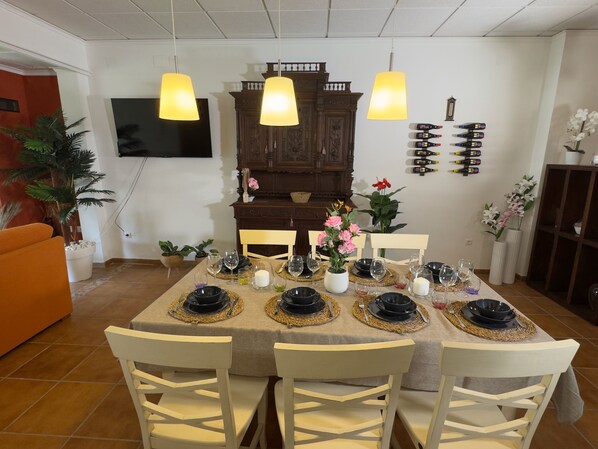 A dining area with a table set for four, surrounded by white chairs, a wooden cabinet, and a television mounted on the wall.