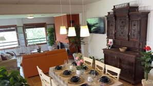 A dining area with a table set for four, surrounded by white chairs, a sofa, and a TV mounted on the wall.