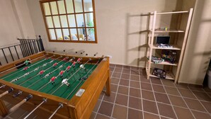 A room with a foosball table, a window with blinds, and a wooden shelf with various items.