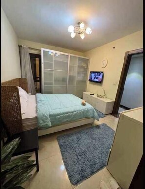 Ein Schlafzimmer mit einem Bett, einem Fernseher, einem kleinen Schreibtisch, einem Fenster mit Vorhängen und einem Deckenventilator.