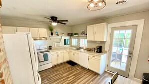 A kitchen with white cabinets, a refrigerator, a stove, and a ceiling fan. - The Little Sandpiper - Charming Port Royal Cottage (Port Royal)
