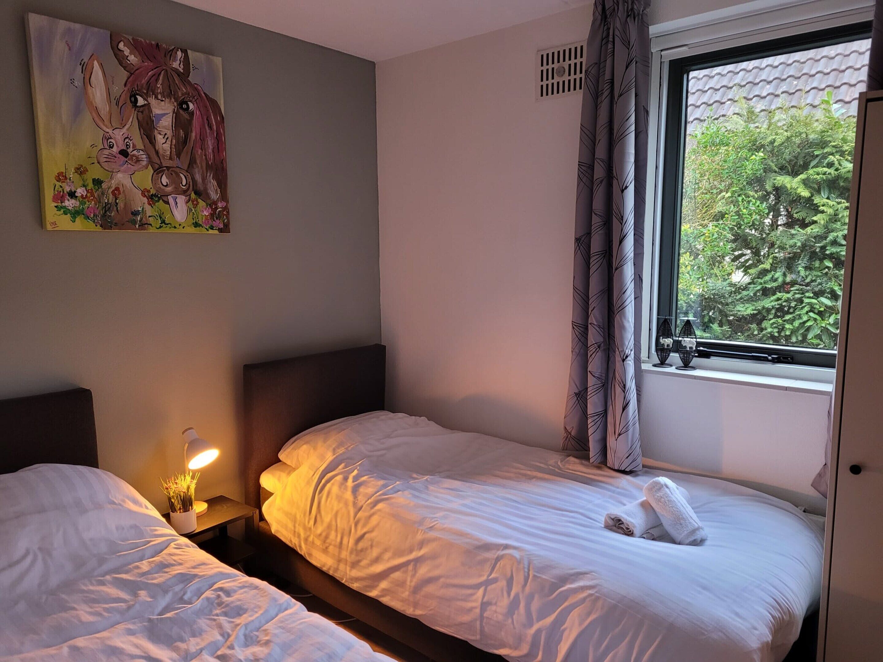 A bedroom with two beds, a nightstand with a lamp, a painting on the wall, and a window with a view of greenery.