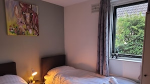 Ein Schlafzimmer mit zwei Betten, einem Nachttisch mit Lampe, einem Bild an der Wand und einem Fenster mit Blick auf Grünflächen.