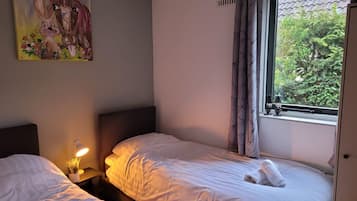 A bedroom with two beds, a nightstand with a lamp, a painting on the wall, and a window with a view of greenery.