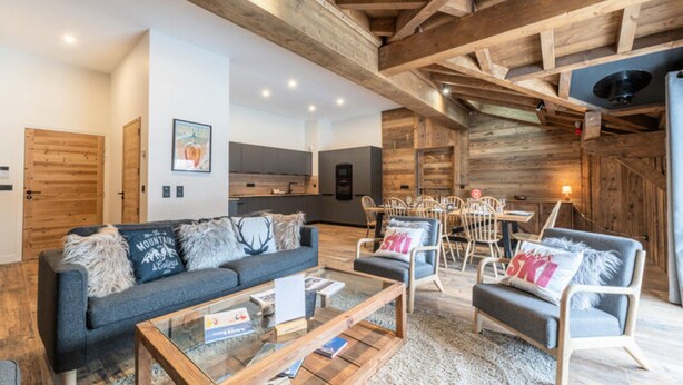 Un salon moderne avec un canapé, des fauteuils et une table basse. Au fond, on aperçoit une cuisine avec un coin repas.