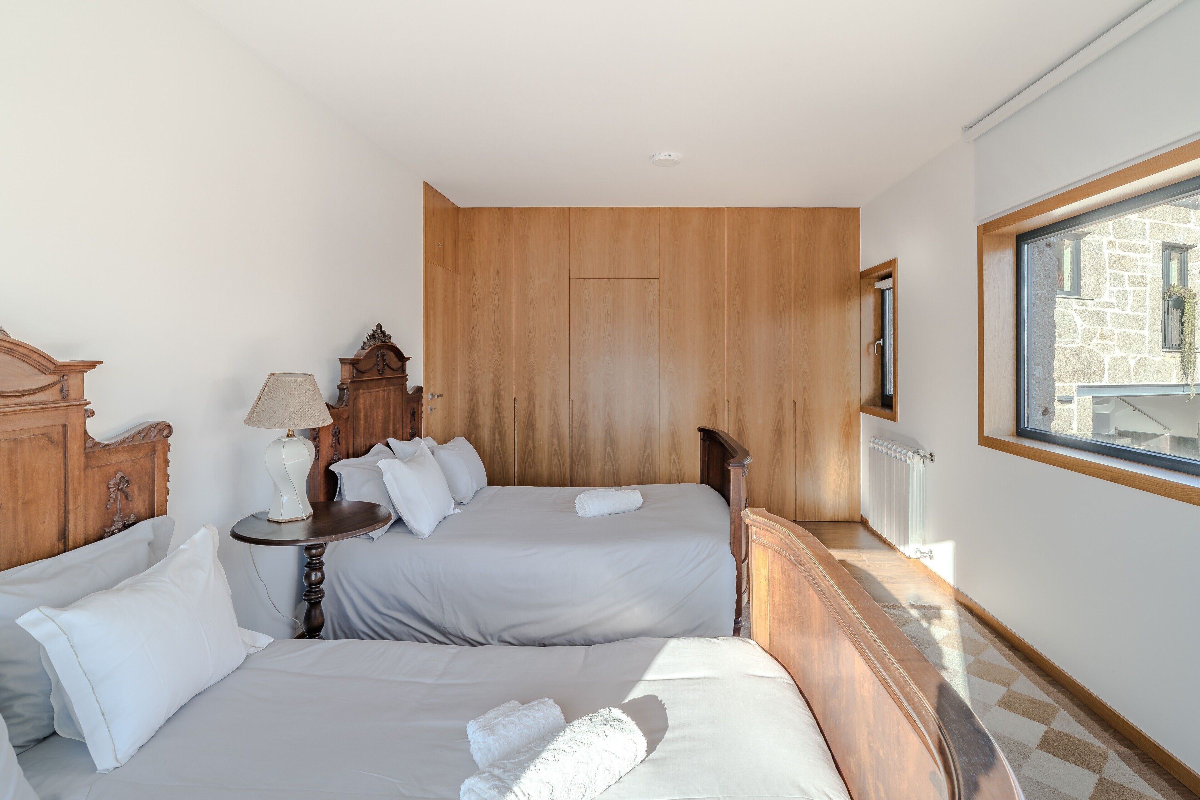 A hotel room with two beds, a wooden headboard, a nightstand, a lamp, and a window with a view of buildings.