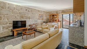 A living room with a stone wall, a flat-screen TV, a sofa, and a wooden cabinet. - Holiday Home 'Casa Feixa Gerês' with Mountain View, Wi-Fi & Air Conditioning (Gerês)