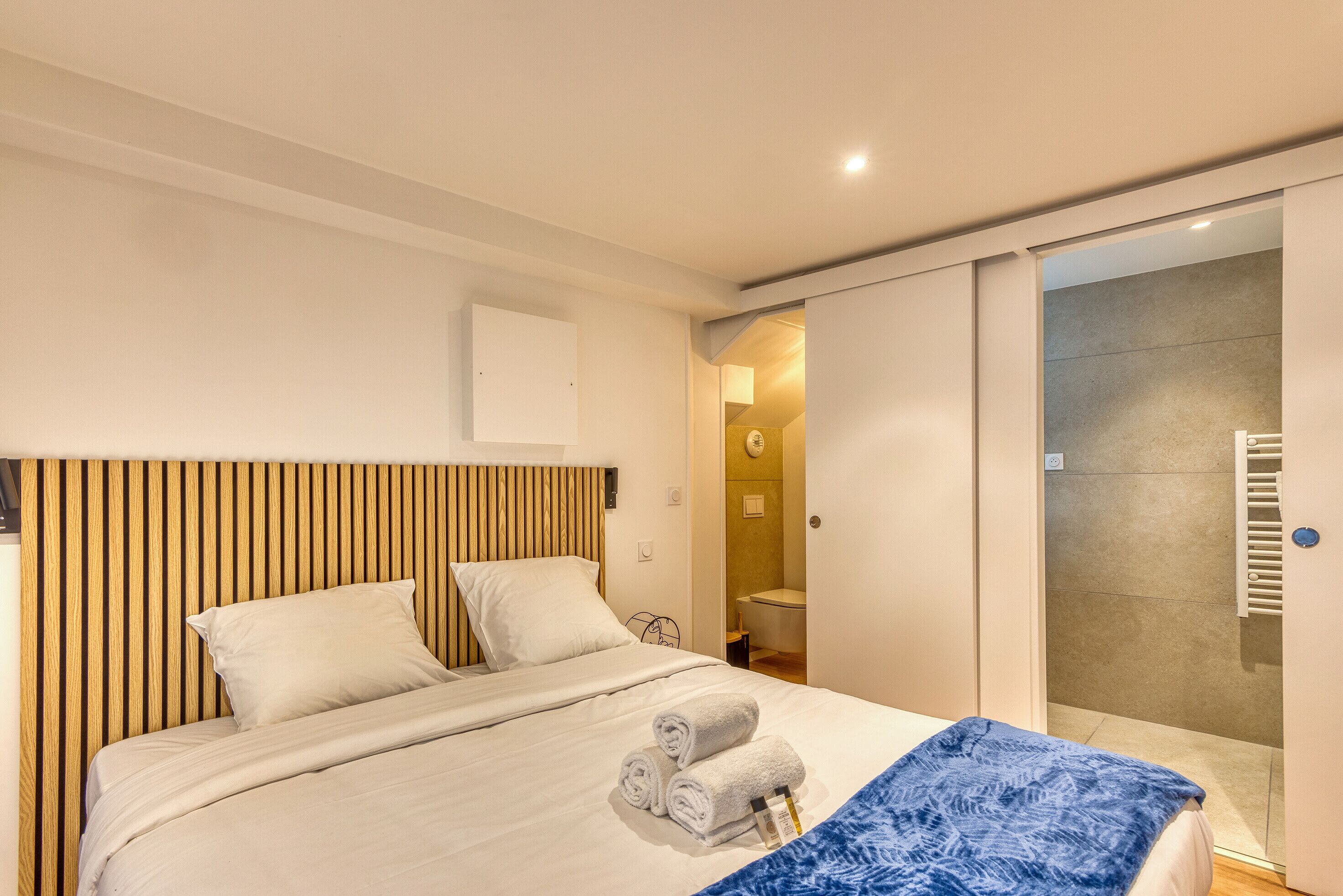 A modern hotel room with a bed, a wooden headboard, a bathroom visible through an open door, and a blue patterned throw blanket on the bed.