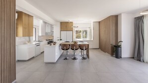 A modern kitchen with white cabinetry, a central island with stools, and a large window providing natural light. - Luna Mare in Agia Marina Chrysochous (Ayia Marina Chrysochous)