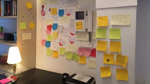 Um quarto com uma parede repleta de anotações e post-its, uma escrivaninha com uma luminária e um telefone.