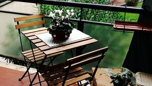Varanda com mesa e cadeiras de madeira, um vaso de plantas e vista para uma área residencial com vegetação e edifícios.