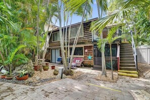 A wooden house with a covered porch, surrounded by tropical plants and a small garden area.