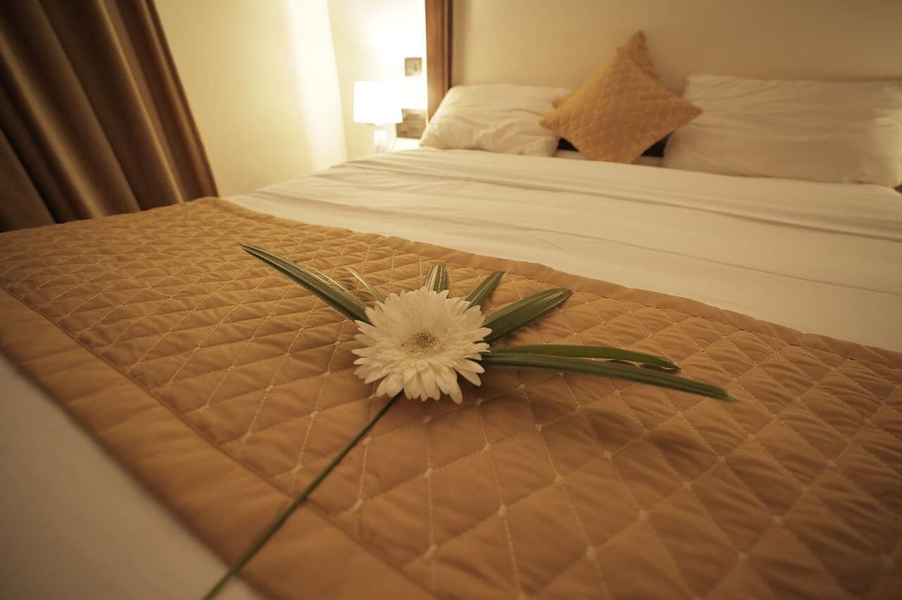A hotel room with a neatly made bed, a quilted brown bedspread, and a single white flower with green leaves on the bed.