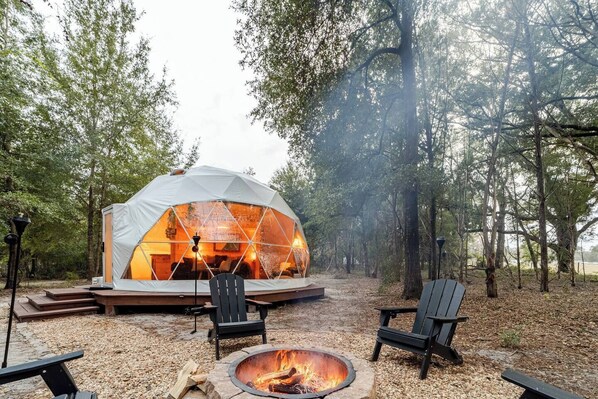 A geodesic dome structure with a fire pit and seating area in a forest setting.