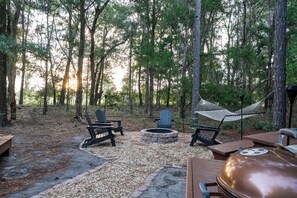 A forested campsite with a fire pit, two Adirondack chairs, a hammock, and a grill.