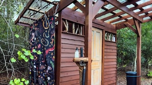 Une cabane en bois avec une véranda, une porte et une fenêtre. On trouve une poubelle noire et un robinet d’eau sur la véranda.