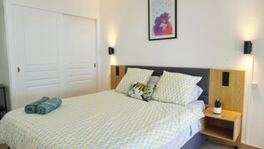 Een net opgemaakt bed met beddengoed met een patroon, een nachtkastje met een plant erop en een ingelijst schilderij aan de muur.