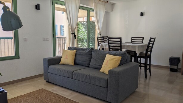 Een woonkamer met een grijze bank, twee gele kussens, een eettafel met stoelen en een raam met gordijnen.