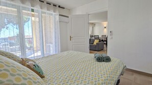 Een slaapkamer met een bed, een raam met gordijnen, een deur en uitzicht op een zithoek met een bank en een tafel.