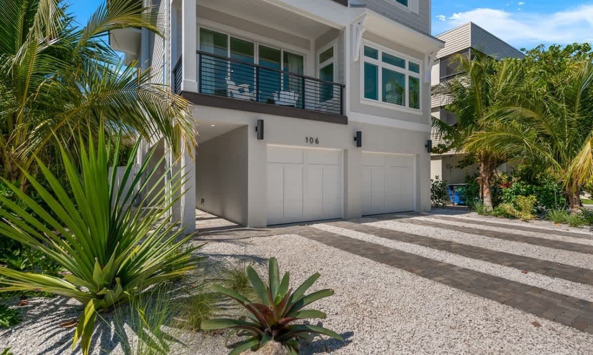 A modern two-story house with a garage, number 106, surrounded by tropical plants.