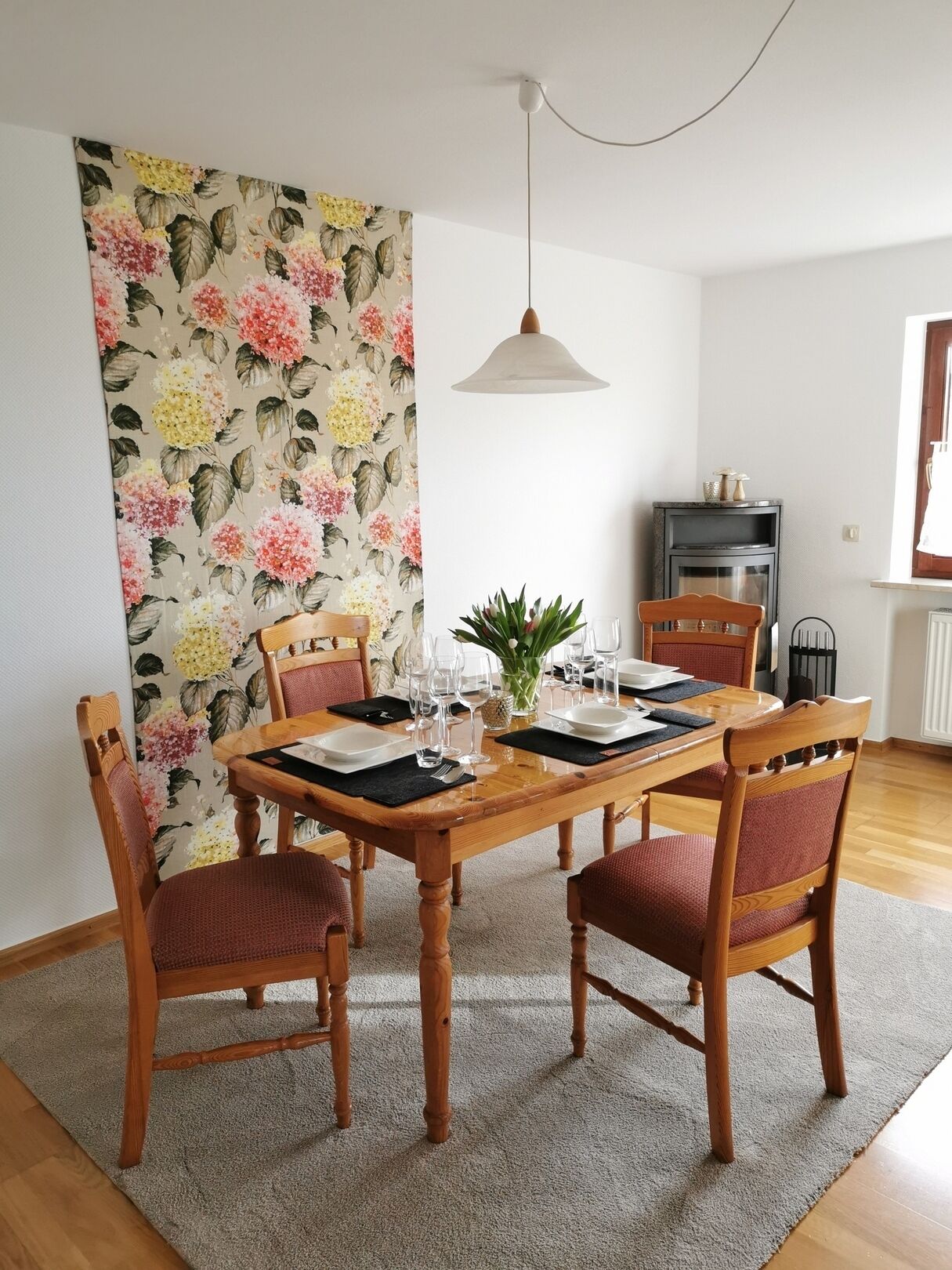 Ein Essbereich mit einem Holztisch, gedeckt für vier Personen, umgeben von Holzstühlen mit roten Kissen. An der Wand hinter dem Tisch ist eine blumige Tapete zu sehen.