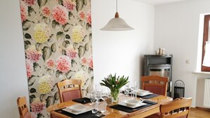 A dining area with a wooden table set for four, surrounded by wooden chairs with red cushions. A floral wallpaper is visible on the wall behind the table. - Vacation apartment "Fröhlich am Baderbichl" (Tutzing)