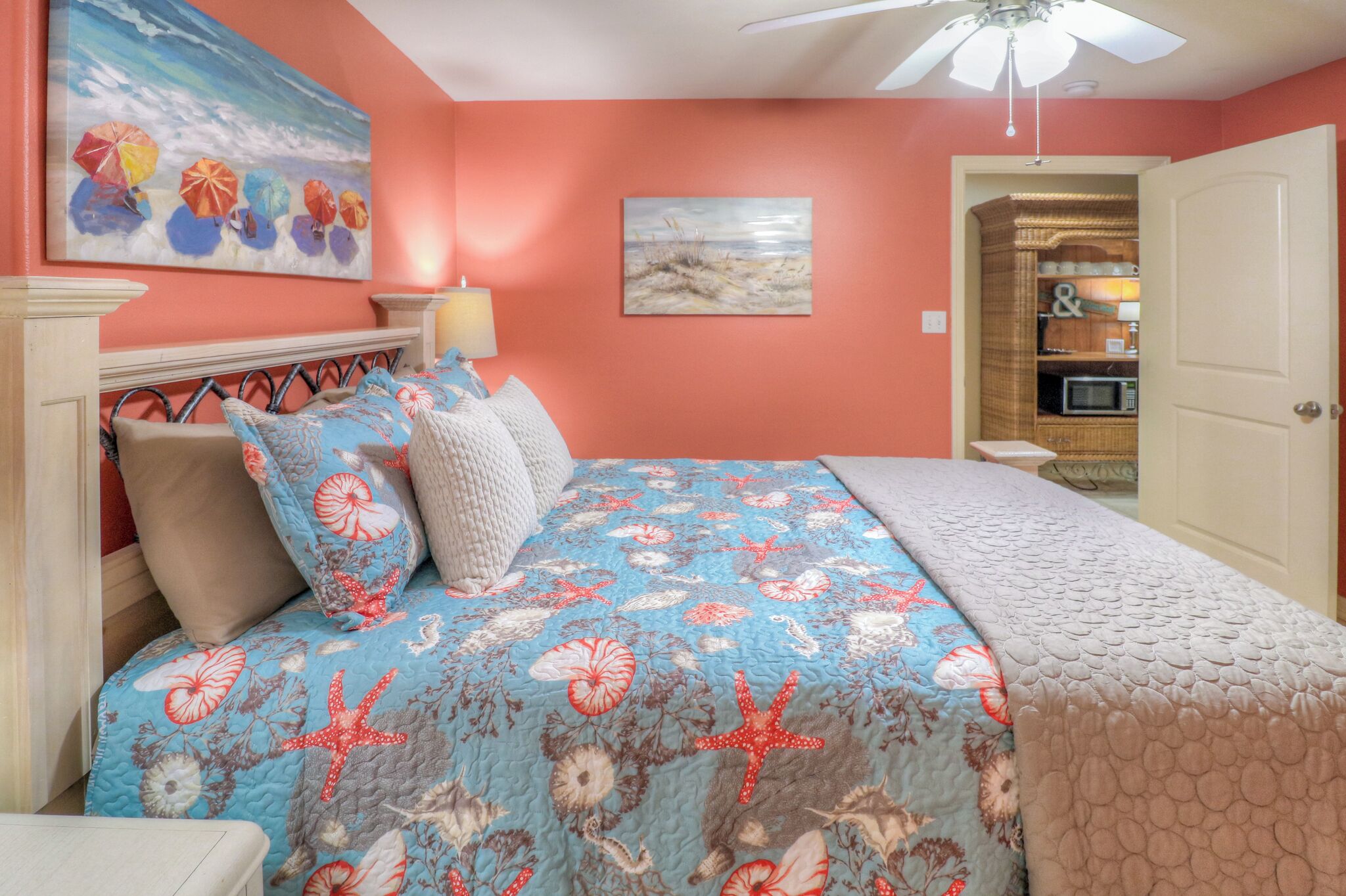 Un dormitorio con cama, ventilador de techo, un cuadro con sombrillas de playa y una puerta que da a una zona de cocina.