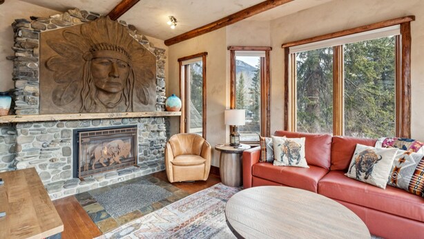 A stone fireplace with a carved head, a red leather sofa, a round wooden coffee table, and large windows with a mountain view.