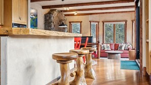 A rustic interior with wooden bar stools, a stone fireplace, a sofa with patterned cushions, and large windows.