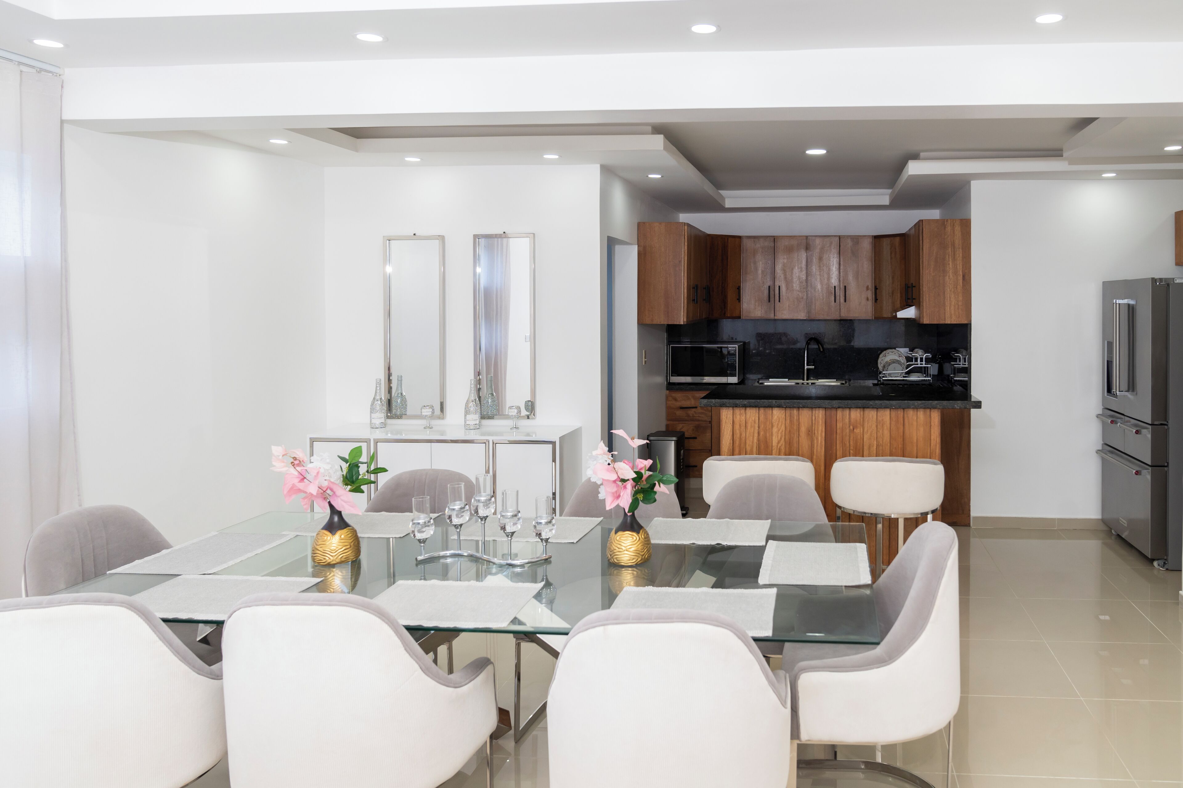 Una cocina moderna con armarios de madera, un refrigerador de acero inoxidable y una zona de comedor con mesa de vidrio y sillas blancas.