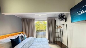 Une chambre à coucher comprenant un lit, un bureau, un portant et une grande fenêtre avec des rideaux.