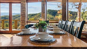 Una mesa de comedor dispuesta con cuencos y platos, rodeada de sillas, con vistas a un paisaje montañoso a través de amplios ventanales.