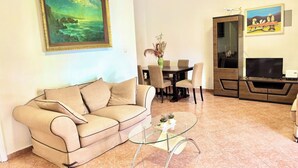 Ein Wohnzimmer mit einem beigefarbenen Sofa, einem Glastisch, einem Esstisch mit Stühlen, einem Fernseher und Kunstwerken an den Wänden.