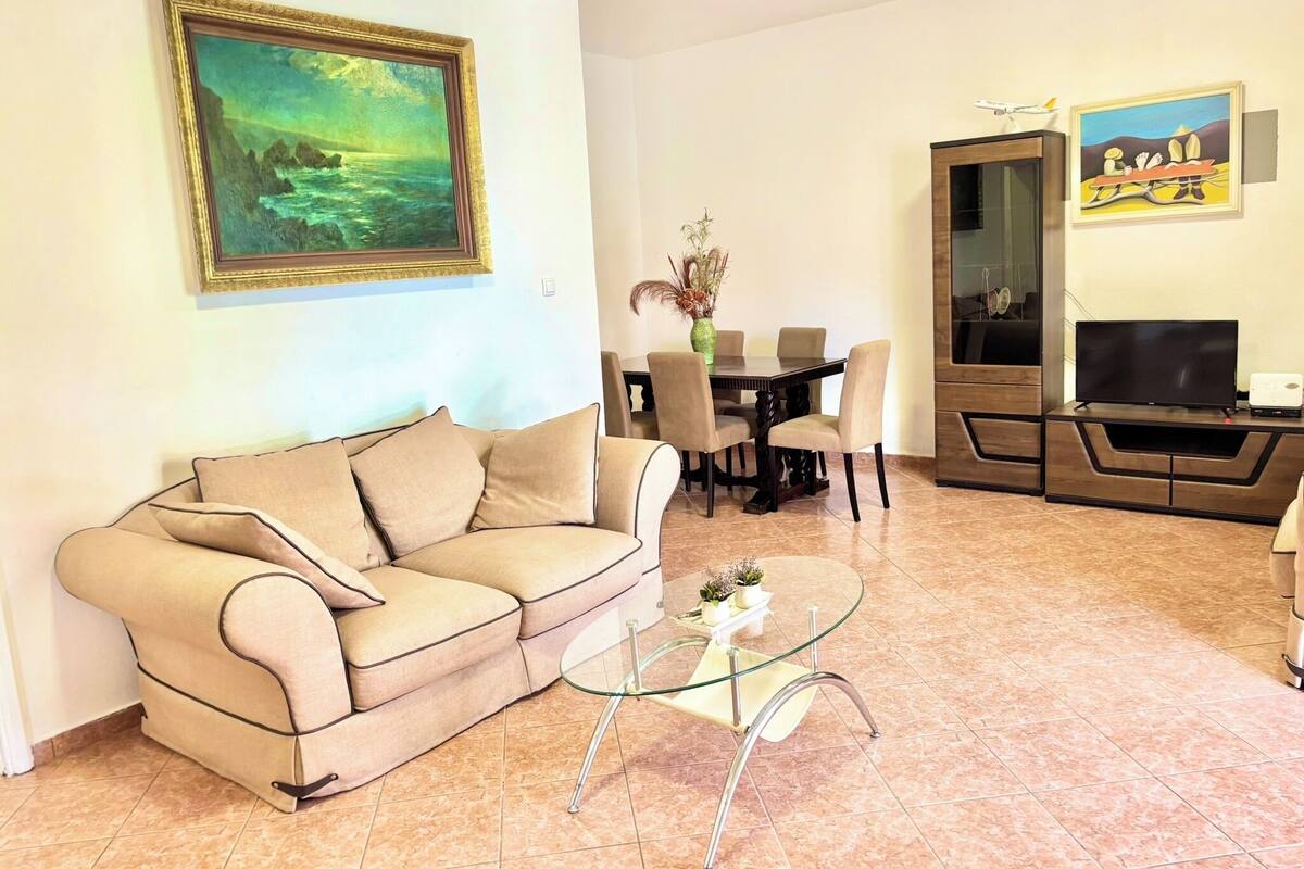 A living room with a beige sofa, a glass coffee table, a dining table with chairs, a TV, and artwork on the walls.