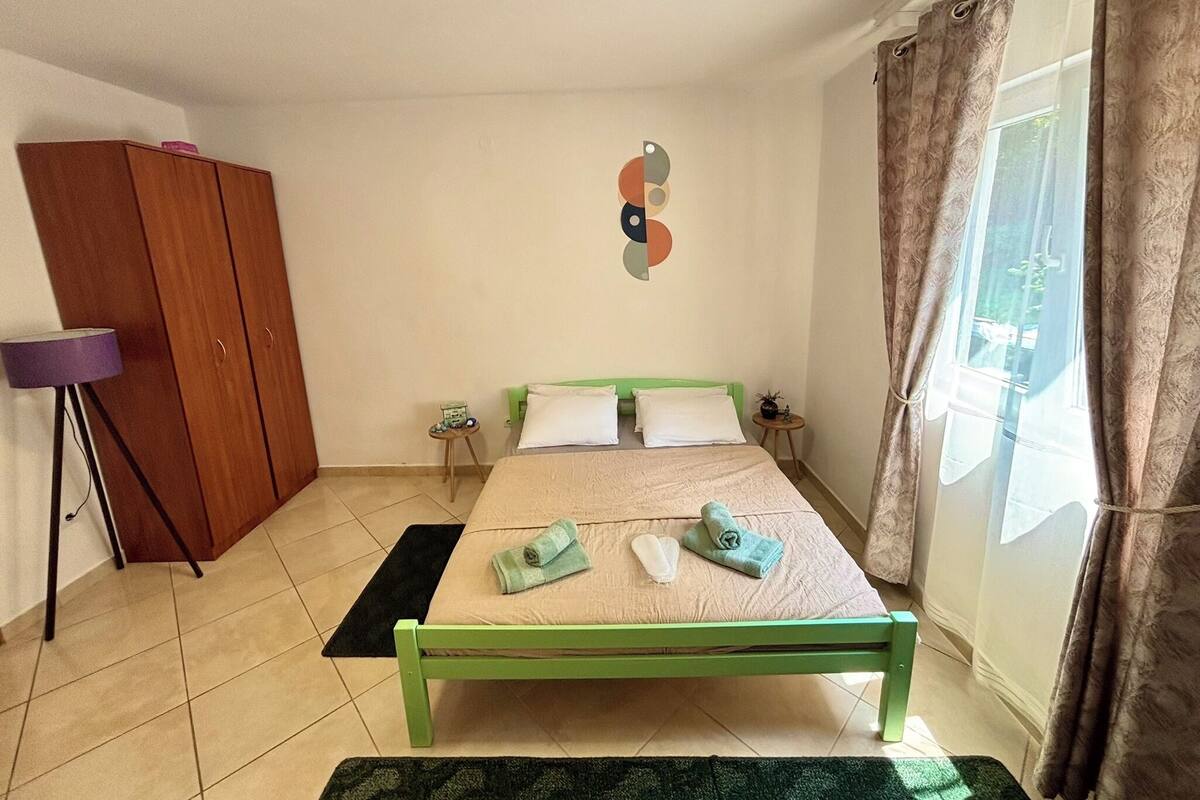 A bedroom with a green bed, a wooden wardrobe, a small table with a lamp, and a window with curtains.