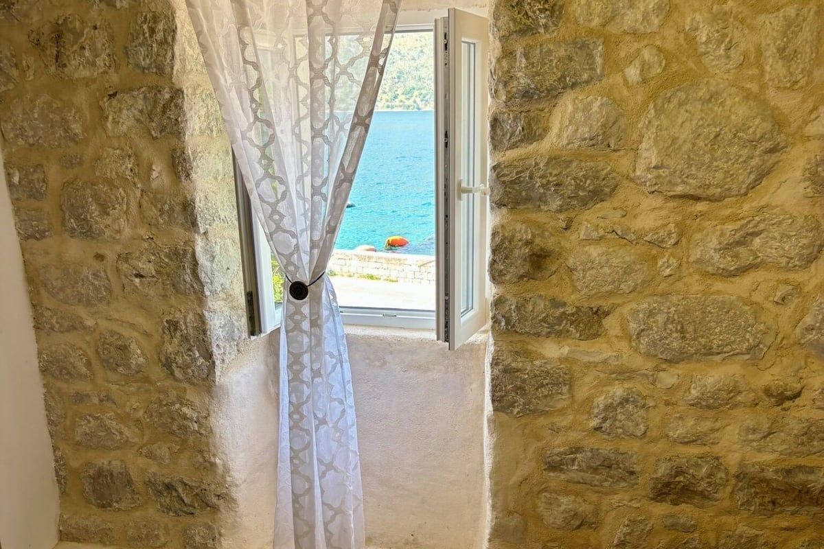 A room with stone walls, a bed with a beige cover, and a window with a view of the sea.