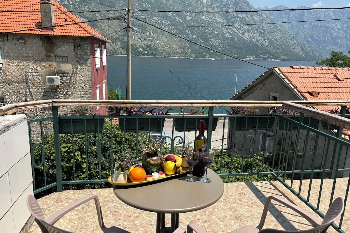 A terrace with a table and chairs, a tray with fruits and wine glasses, and a view of a lake and mountains.